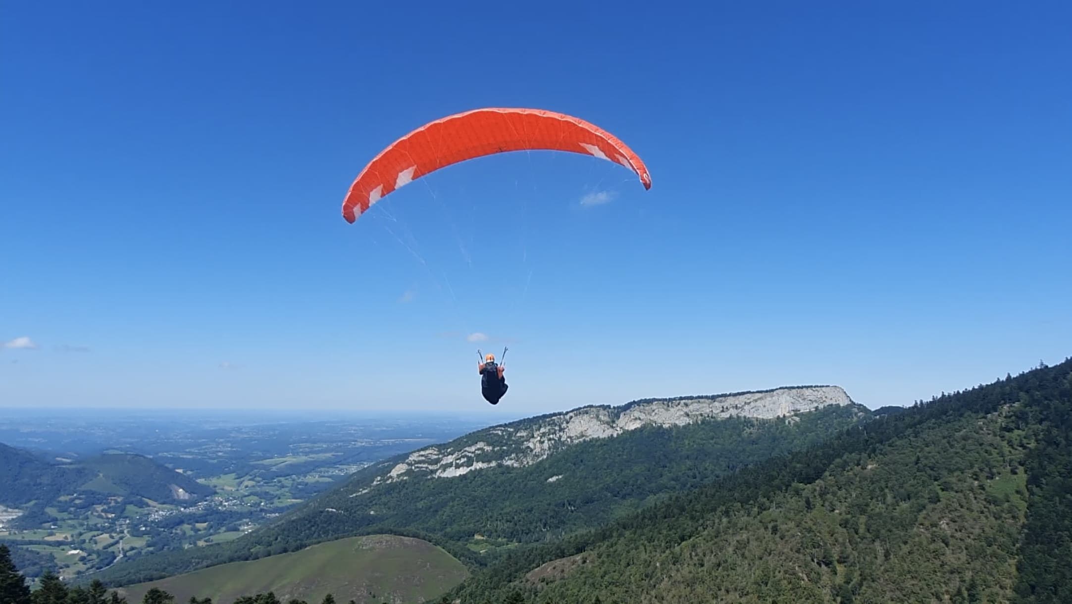 Parapente orange en vol dans la vallée d'Aure, vue dégagée sur la plaine.